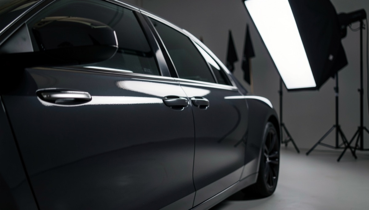 Portière de voiture sombre filmée de côté en studio, long reflet blanc d'une grande boîte à lumière qui court sur la tôle, drapeaux noirs flous au fond.